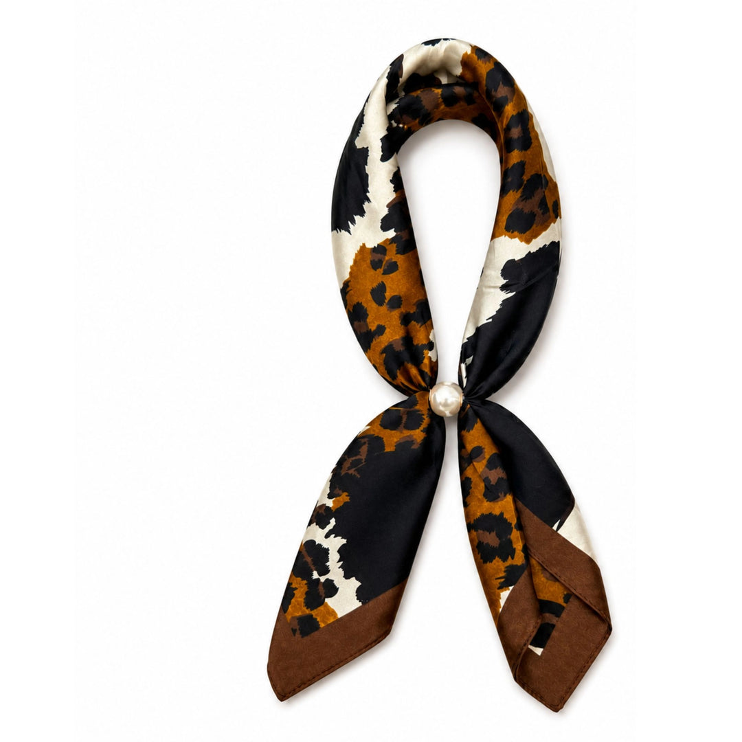 A silk scarf with a cow-print pattern in brown, black, and cream, tied in a loop with a pearl accent at the center, on a solid white background.