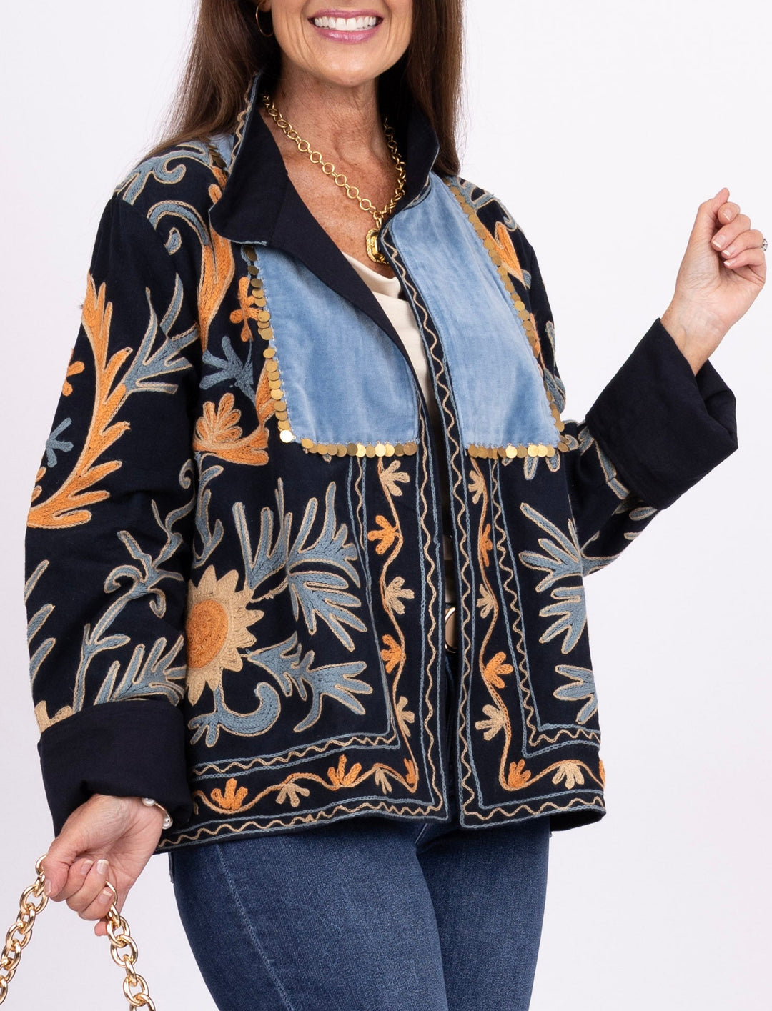 A woman modeling a black embroidered jacket with orange, blue, and gray botanical patterns, paired with blue jeans, brown boots, and holding a blue woven handbag with a gold chain strap against a white studio background.