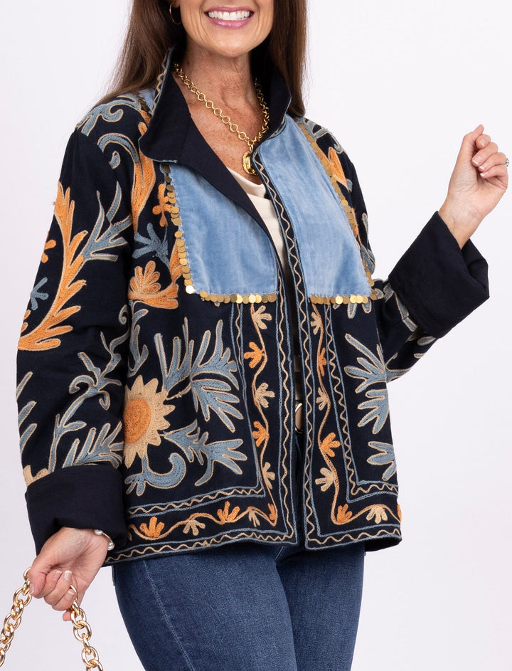A woman modeling a black embroidered jacket with orange, blue, and gray botanical patterns, paired with blue jeans, brown boots, and holding a blue woven handbag with a gold chain strap against a white studio background.