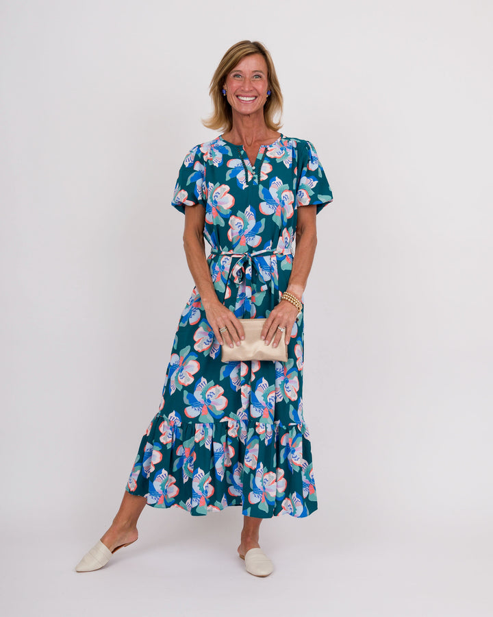 A woman modeling a short-sleeve midi dress with a ruffled tiered hem, featuring a teal green base with a pink, blue, and white floral pattern. She is holding a small beige clutch and wearing beige pointed-toe flats, standing against a plain light gray studio background.