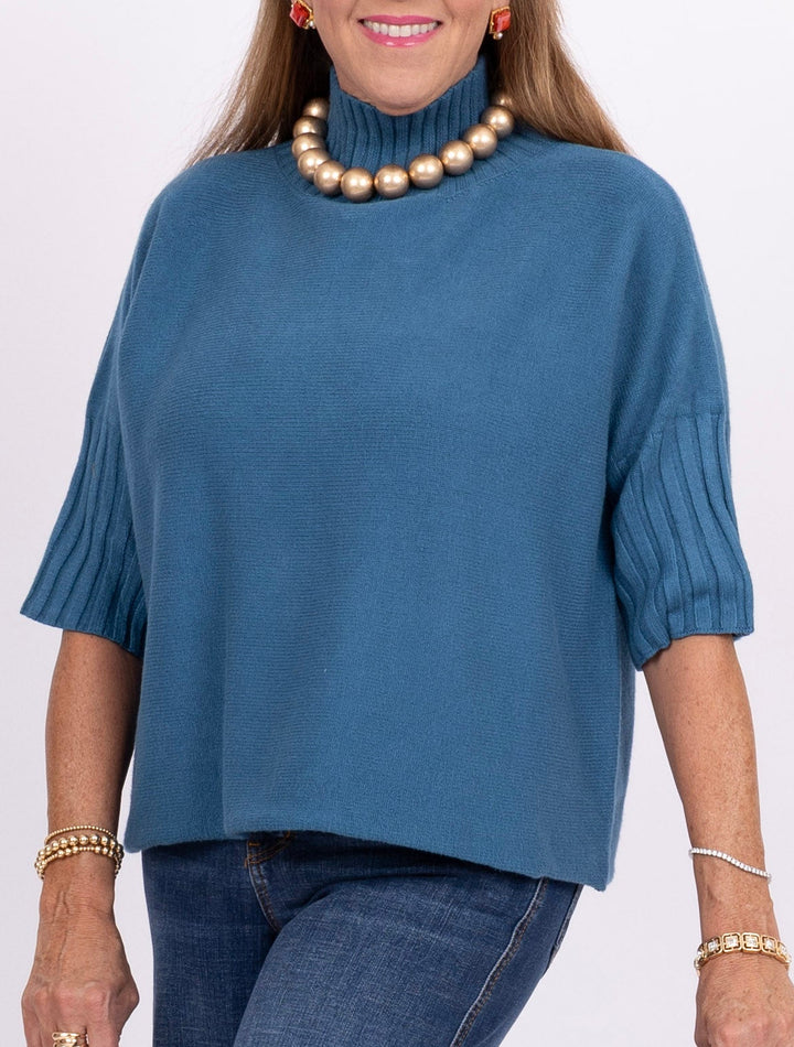 A woman wearing a slate blue turtleneck sweater with ribbed elbow-length sleeves, paired with blue jeans and gold jewelry, against a plain white background.