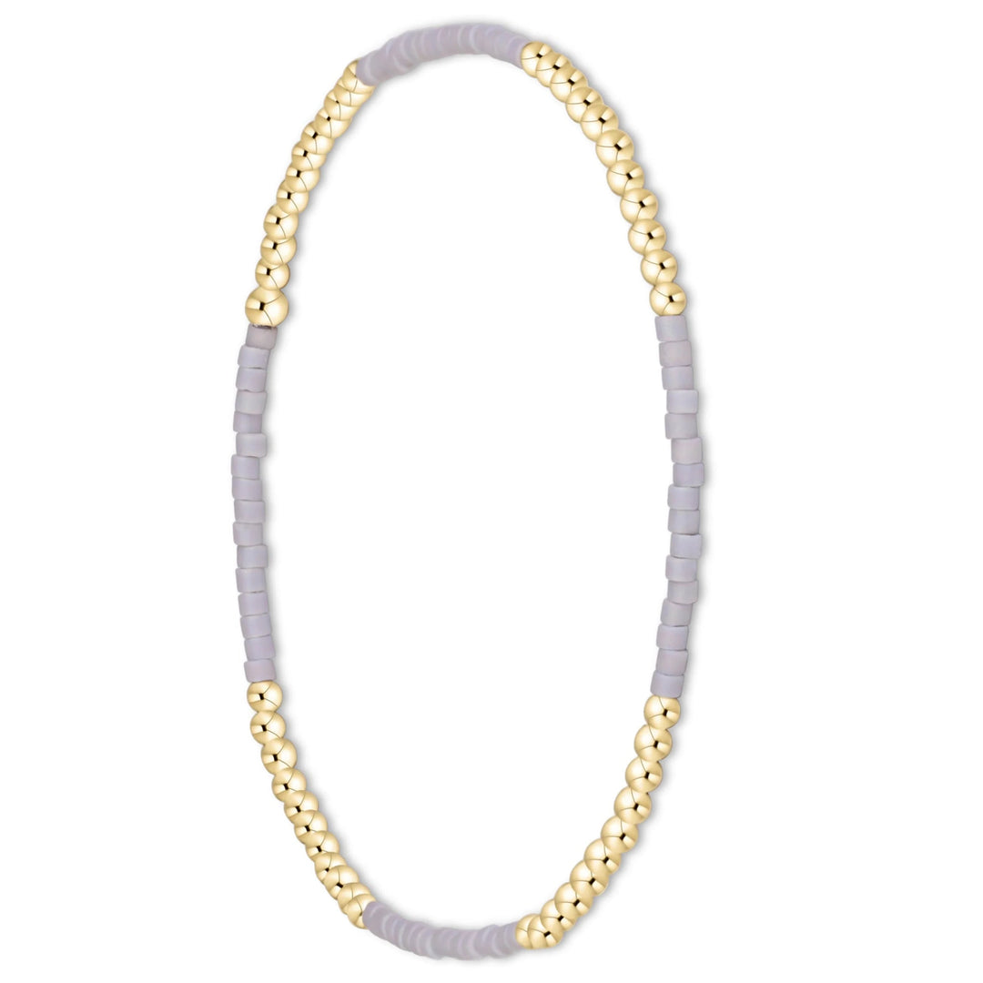 A delicate beaded stretch bracelet featuring alternating strands of matte light purple-gray cylindrical beads and polished gold-tone round beads, shown on a plain white background.