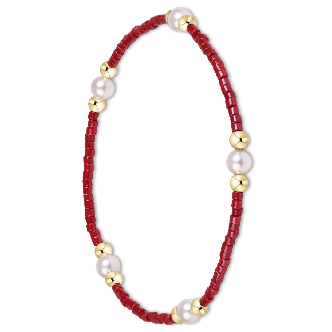 A delicate beaded bracelet featuring deep red cylindrical beads, polished white pearls, and shiny gold spacer beads, arranged in a circular shape on a solid white background.
