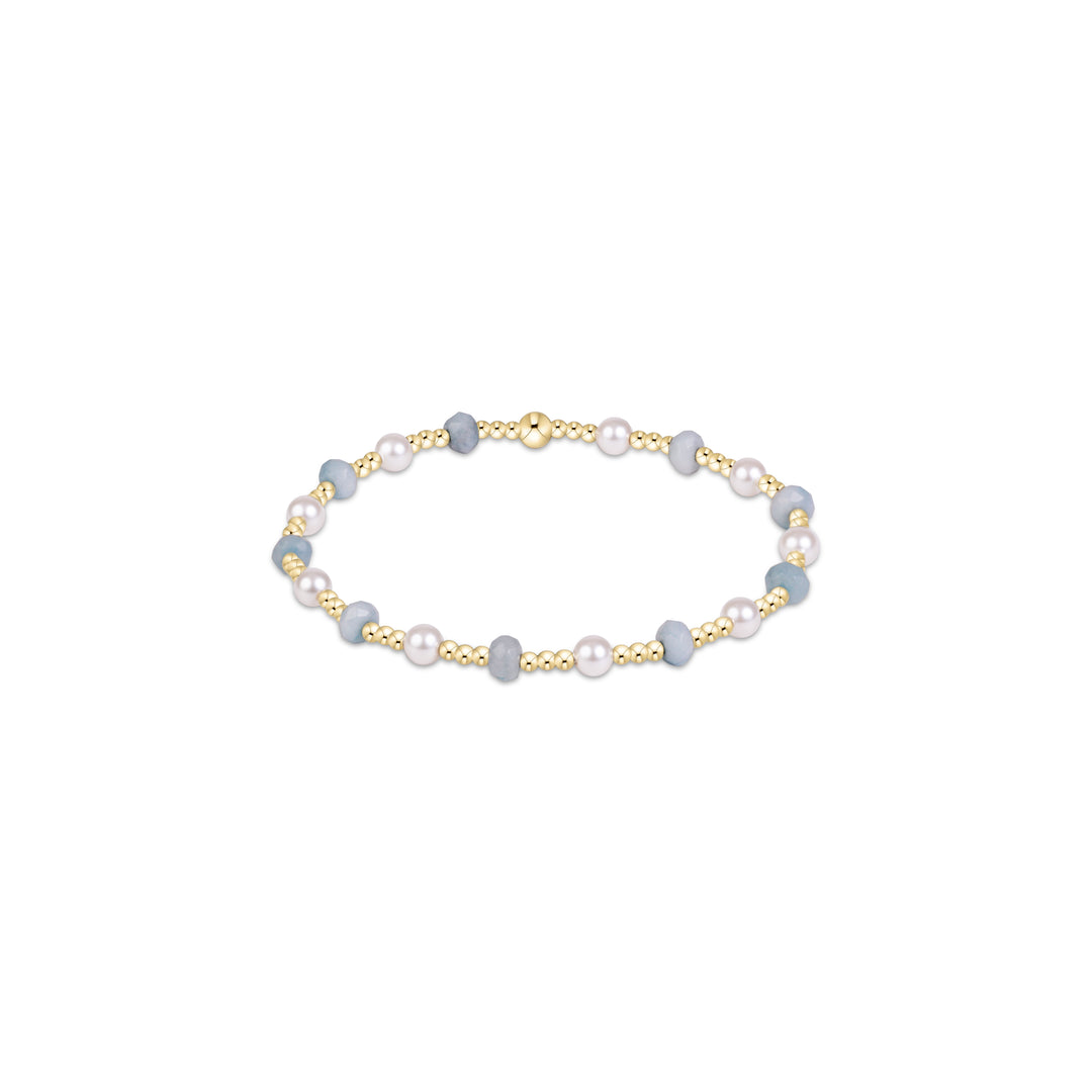 A delicate beaded bracelet featuring light blue-gray beads, white pearls, and polished gold-tone spacer beads, arranged in an oval shape on a clean white background.
