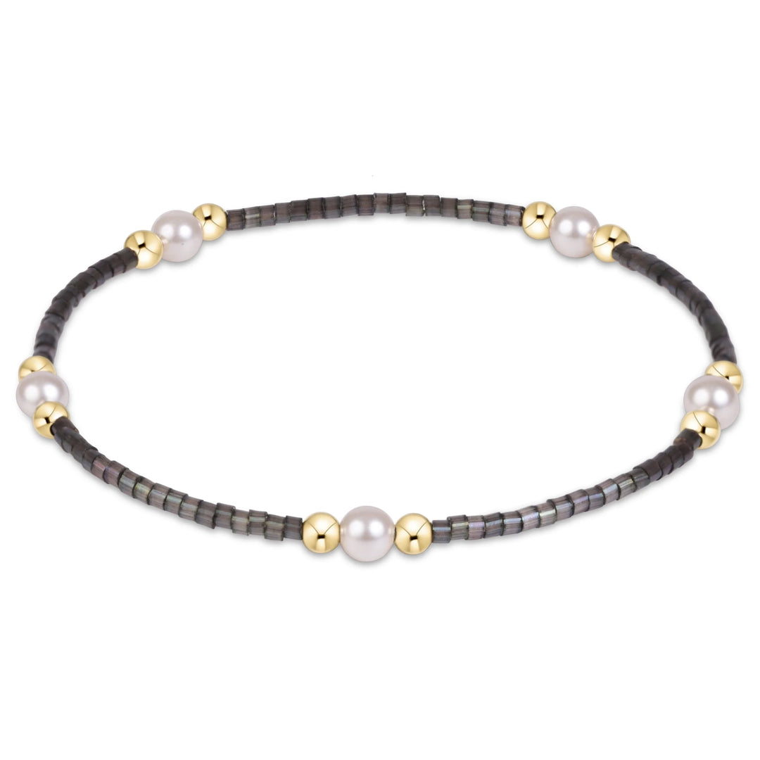 A delicate beaded bracelet featuring dark gray faceted beads, three large white pearls, and three polished gold spacer beads, arranged in an oval shape on a white background.