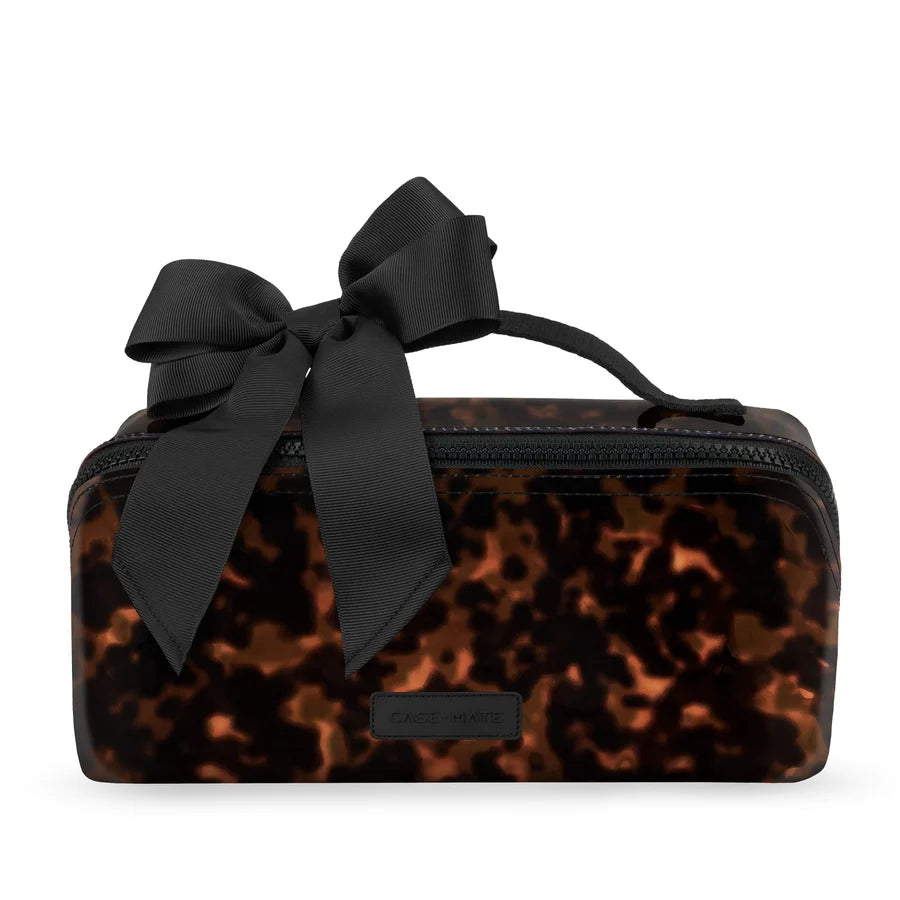 A rectangular cosmetic pouch with a glossy tortoiseshell pattern in brown and black, featuring a large black satin bow on top and a small black brand plaque that reads &