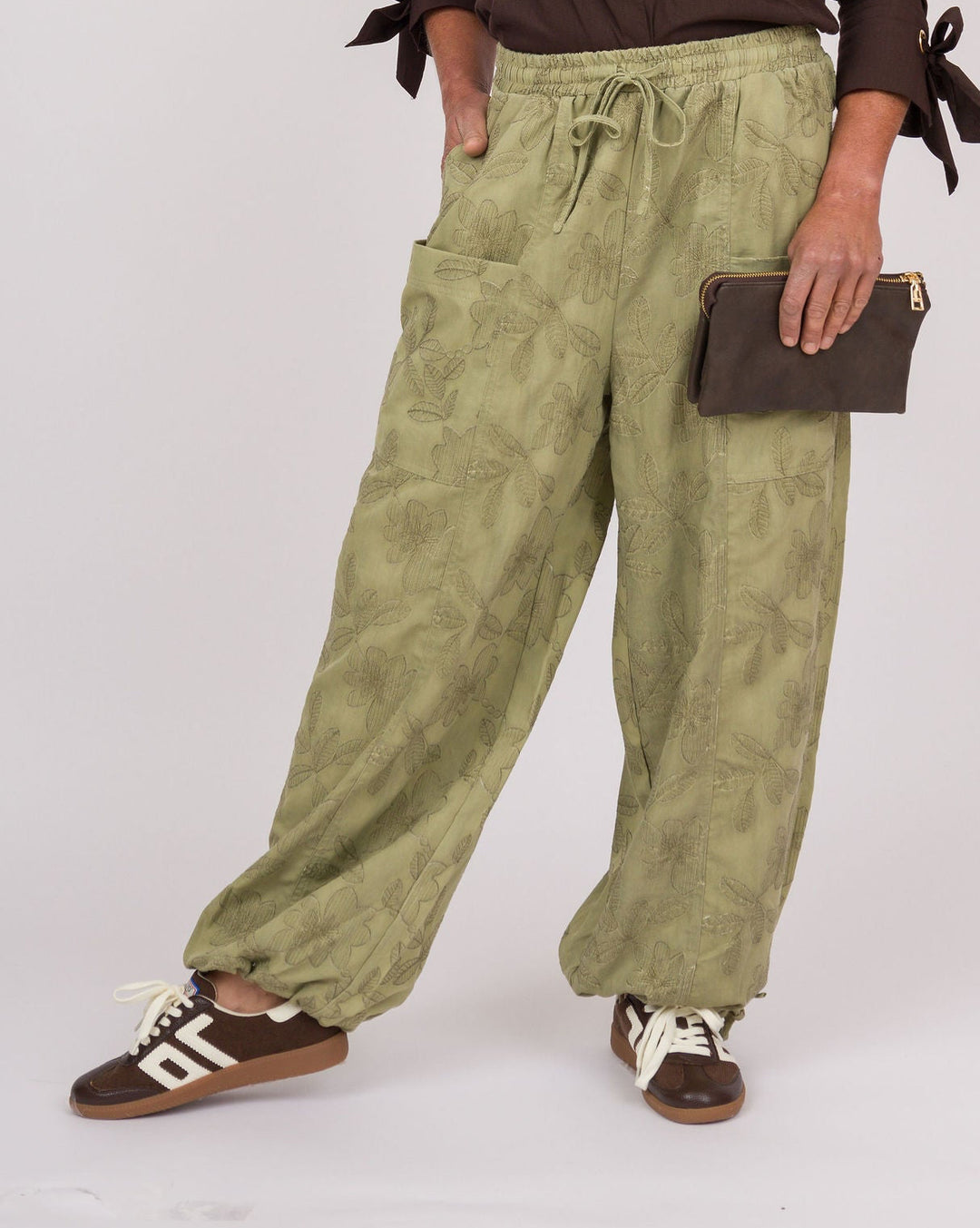 A woman modeling a dark brown button-up shirt with tie details on the sleeves, paired with wide-leg olive green patterned pants, brown sneakers, and holding a small brown clutch against a light gray studio background.