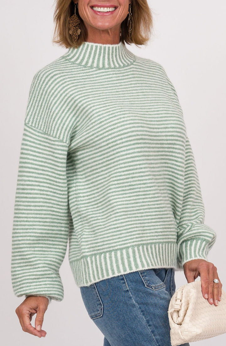 A woman modeling a light green and white striped mock neck sweater, blue jeans, and beige shoes, holding a cream clutch bag against a light gray studio background.