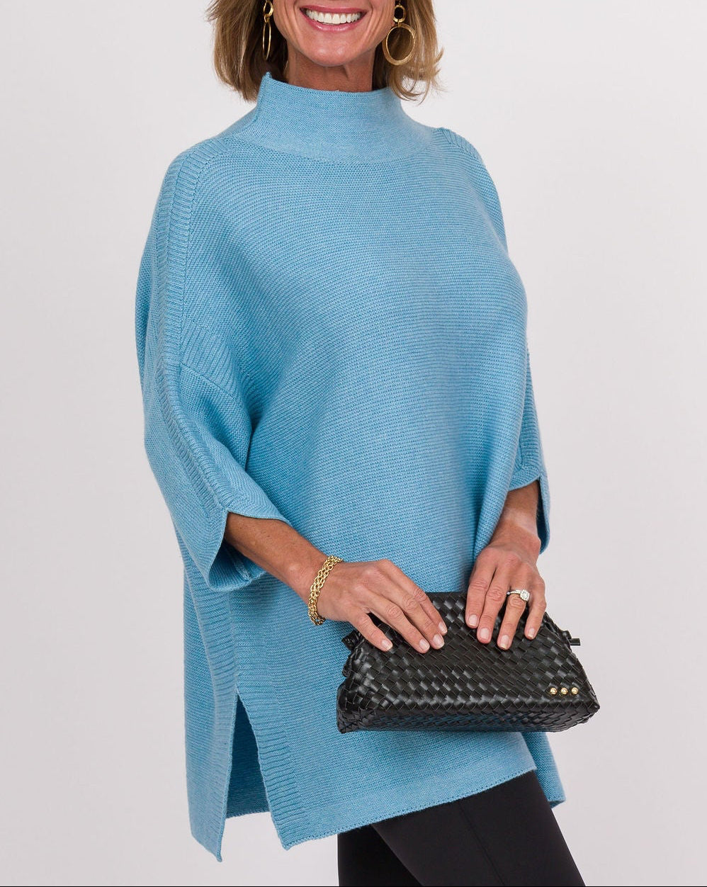 A woman modeling a light blue knit poncho-style top with black leggings and white sneakers, holding a black clutch bag against a plain light gray studio background.