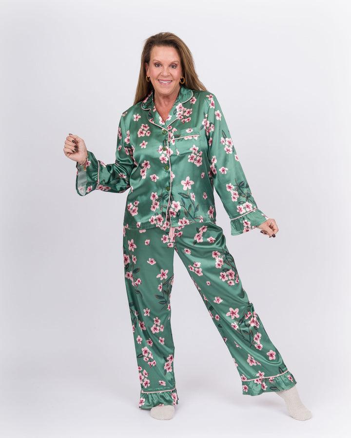 A woman modeling a two-piece satin pajama set with a green base and pink floral print, standing against a light gray studio background.