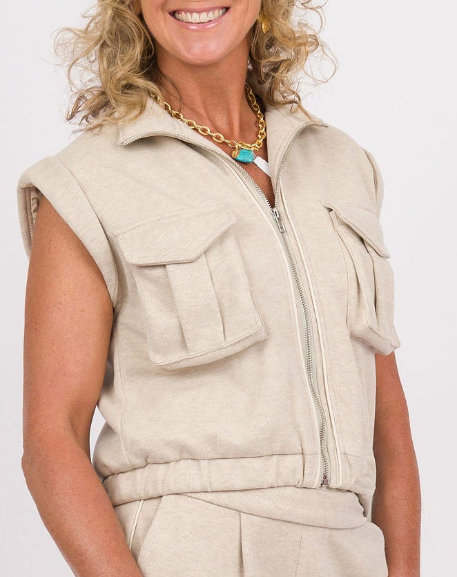A woman modeling a light beige linen utility jumpsuit with a sleeveless vest-style top, wide-leg pants, and a matching belt, holding a small brown leather handbag against a white studio background.