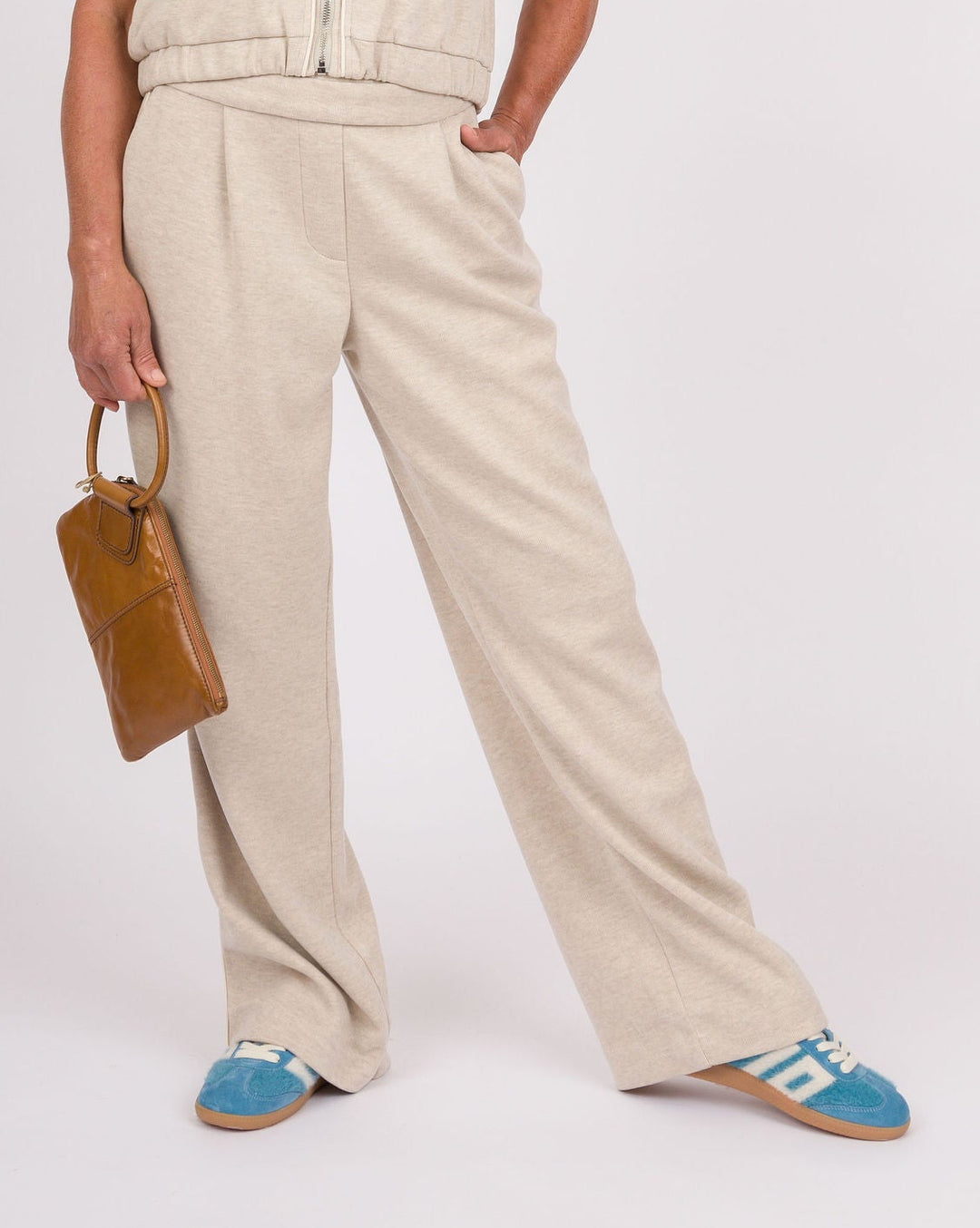 A woman modeling a matching beige two-piece set consisting of a sleeveless zip-front vest with front flap pockets and wide-leg trousers, styled with blue sneakers, a turquoise necklace, and a brown handbag against a white studio background.