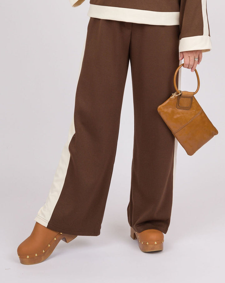 A woman modeling a brown two-piece lounge set with cream contrast trim, featuring a long-sleeve top with a wide collar and matching wide-leg pants. She is holding a small brown handbag against a plain light gray studio background.