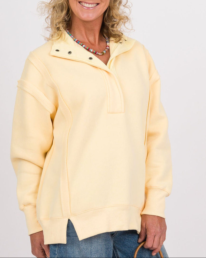 A smiling woman stands against a light gray studio background wearing a pale yellow half-zip pullover, loose blue jeans, white sneakers, and holding a small tan leather handbag.