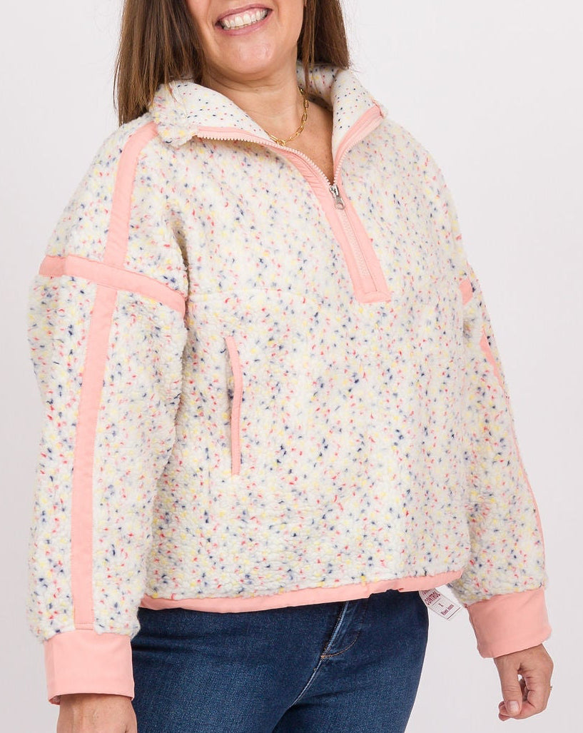 A woman modeling a cream-colored quarter-zip fleece jacket with pink trim and a subtle floral pattern, paired with blue jeans, blue sneakers, and holding a bright pink handbag against a light gray studio background.