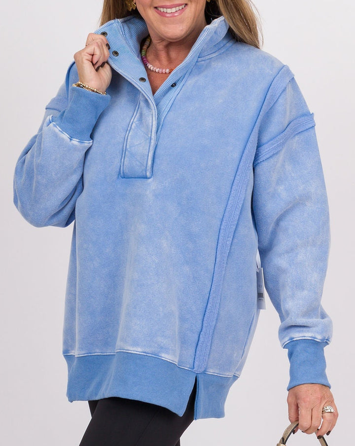 A woman modeling an oversized light blue half-zip pullover sweatshirt, paired with black pants and white sneakers, while holding a small beige handbag against a plain light gray studio background.