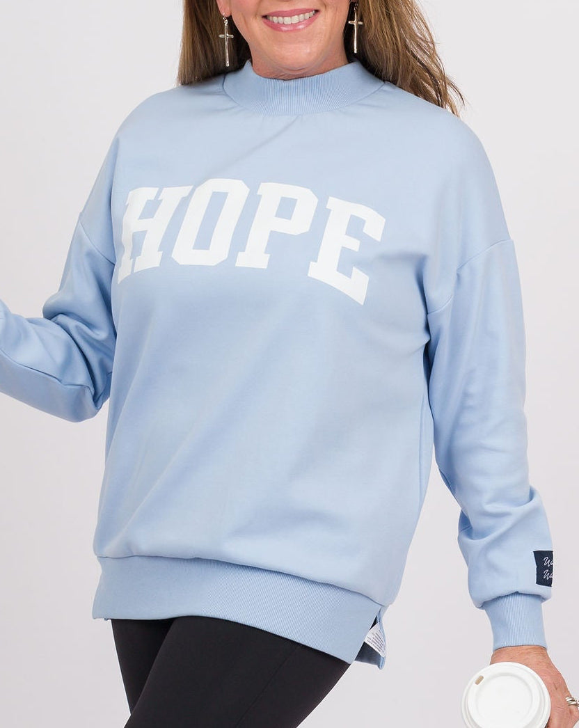 A woman modeling a light blue crewneck sweatshirt with 'HOPE' printed in white arched letters across the chest, paired with black leggings and beige sneakers, holding a white coffee cup against a light gray studio background.