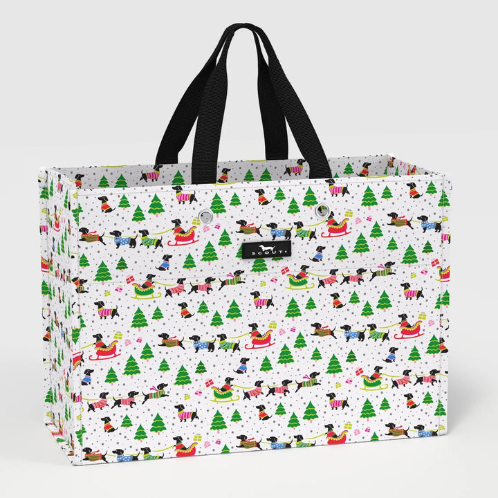 A large tote bag with black handles, featuring a white background covered in a festive Christmas pattern of dogs in sleds, green Christmas trees, and snowflakes. A black leather brand tag for &