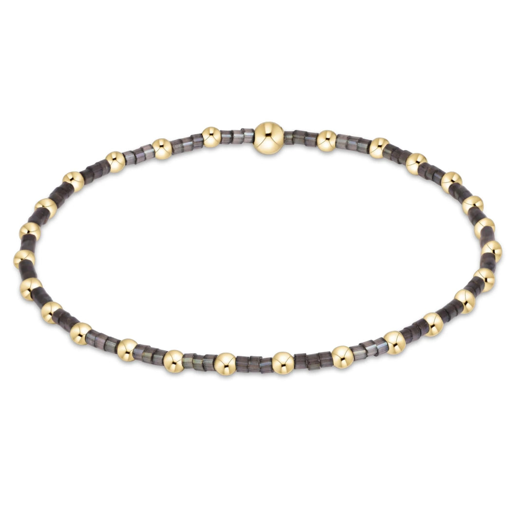 A beaded bracelet featuring alternating dark gray and metallic gold beads, with a larger polished gold bead at the center, displayed on a clean white background.