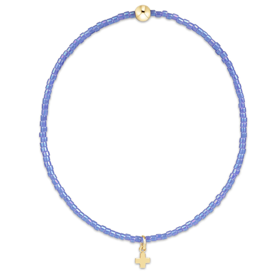 A delicate beaded necklace in light periwinkle blue, featuring a small gold cross pendant and a gold clasp, displayed on a clean white background.