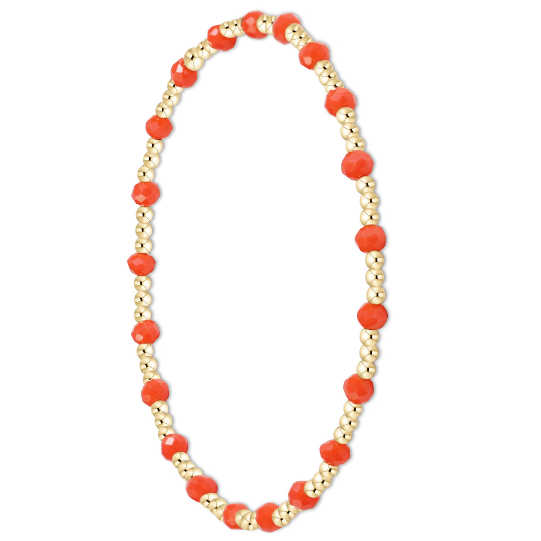 A beaded stretch bracelet featuring faceted orange-red coral beads and small metallic gold spacer beads, arranged in an oval shape on a plain white background.