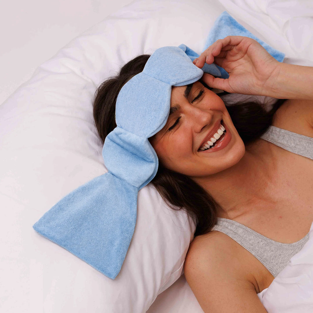 A woman lying in bed with a light blue eye mask on her forehead, smiling and touching the mask. She is wearing a gray tank top and white bedding.