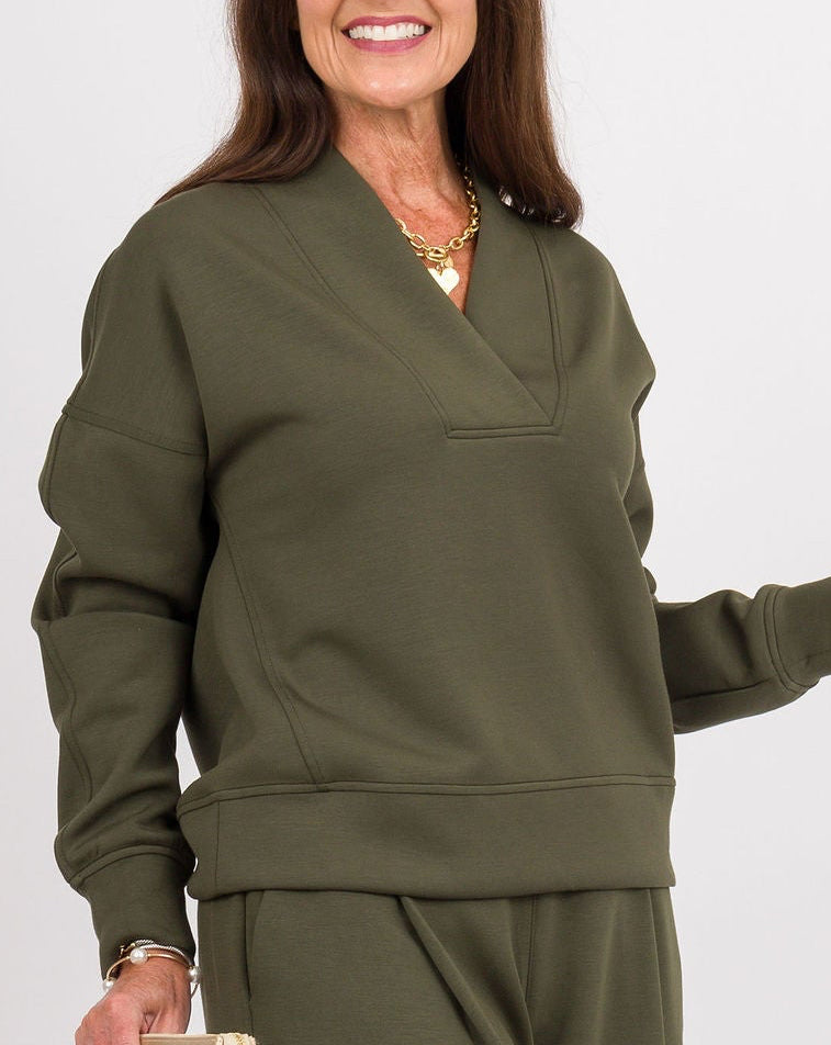 A woman modeling an olive green lounge set consisting of a long-sleeve V-neck top and matching wide-leg pants, standing against a light gray studio background.