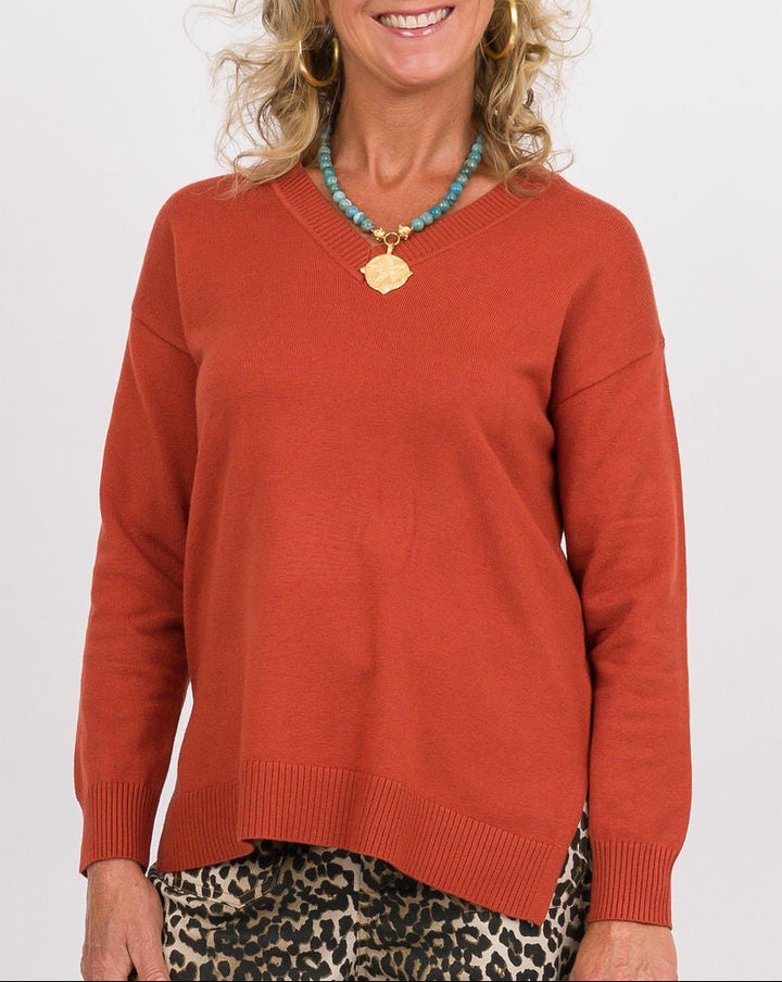 A smiling woman stands against a light gray studio background wearing a rust-orange long-sleeve top, wide-leg leopard-print pants, and dark brown slide sandals. She is holding a dark brown clutch.