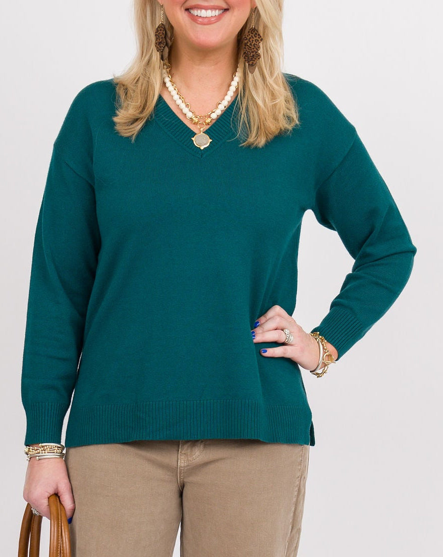 A woman modeling a dark teal green long-sleeve knit sweater, paired with beige wide-leg trousers, brown clogs, and a brown leather handbag, against a plain light gray studio background.