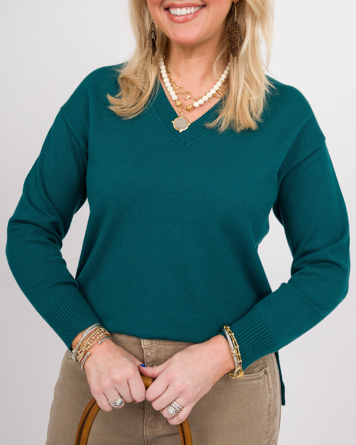 A woman wearing a teal green V-neck knit sweater, styled with khaki pants, layered gold necklaces, bracelets, and rings, against a light gray studio background.