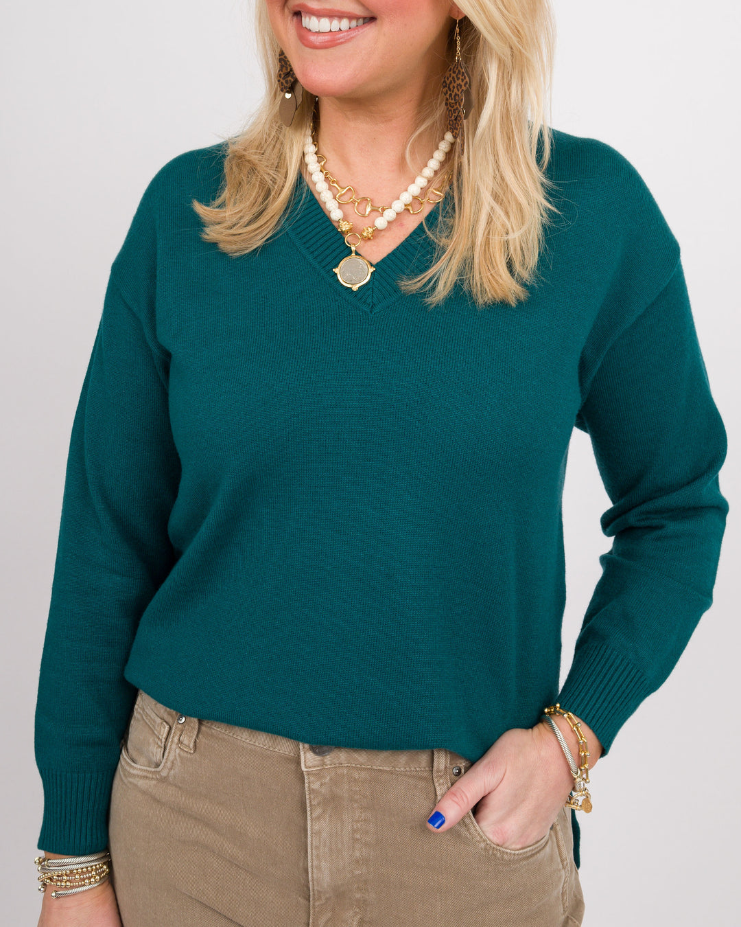 A woman wearing a dark teal green V-neck knit sweater, styled with khaki pants, layered necklaces, and gold bracelets against a light gray background.