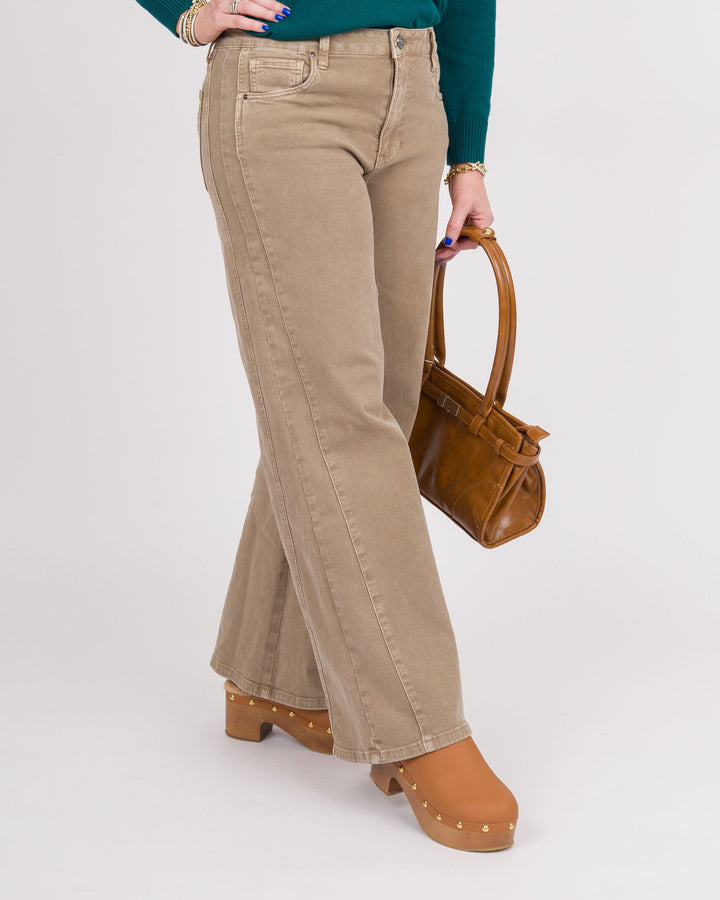 A person wearing beige wide-leg jeans, a teal long-sleeve top, tan clogs, and holding a brown handbag against a light gray studio background.