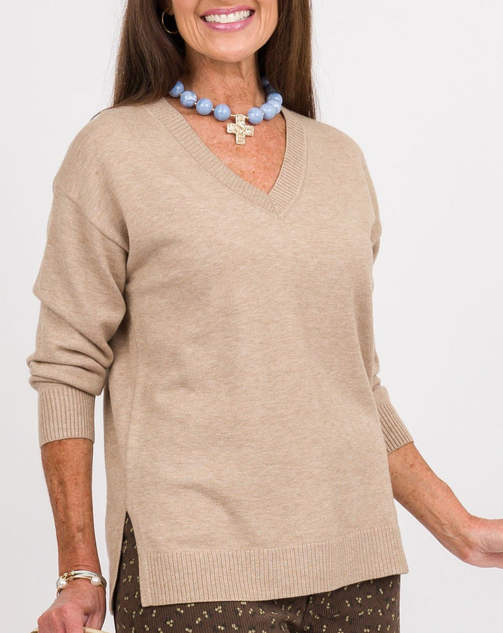 A woman modeling a beige V-neck knit sweater and brown wide-leg pants with a small white pattern, styled with a gold clutch, a blue beaded necklace, and metallic loafers against a plain white studio background.