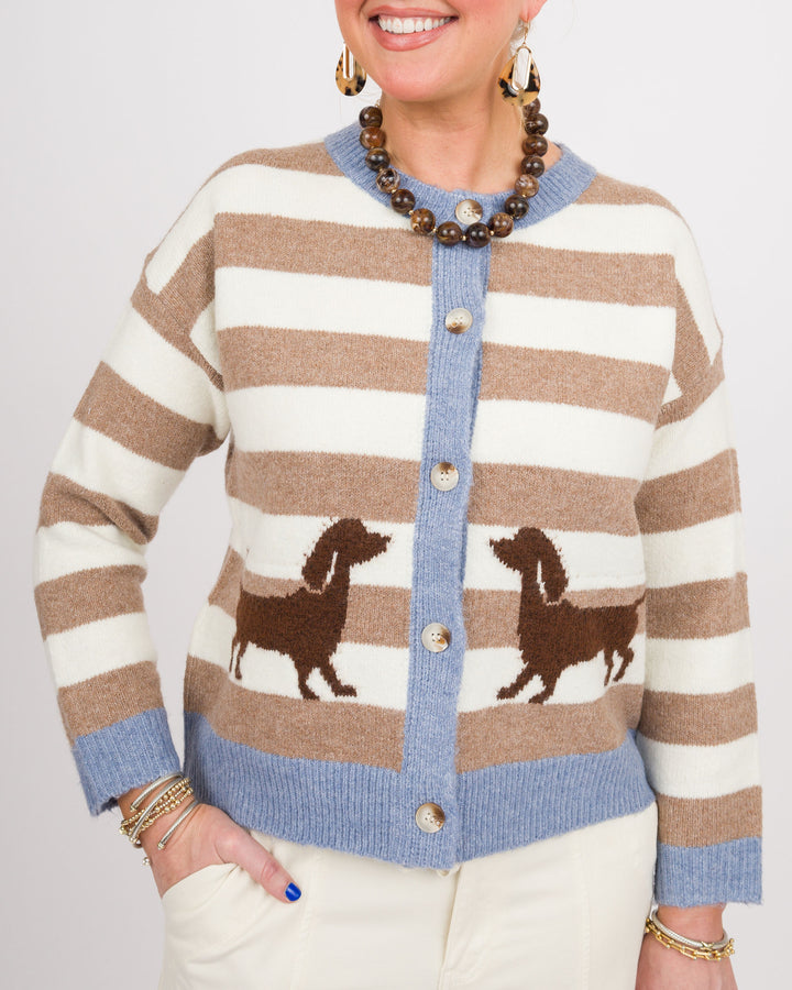 A woman wearing a beige and white striped cardigan with brown dachshund motifs and a light blue ribbed trim, paired with cream pants and gold jewelry against a light gray background.