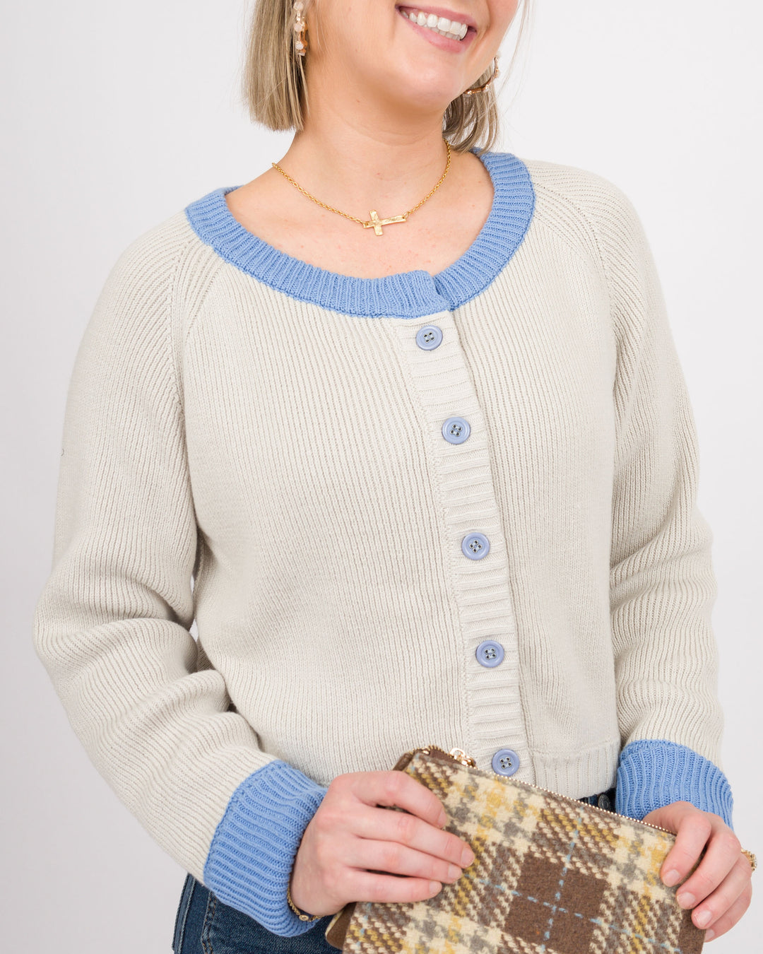 A woman wearing a cream ribbed cardigan with light blue trim and buttons, holding a brown and beige plaid clutch purse against a plain light gray background.
