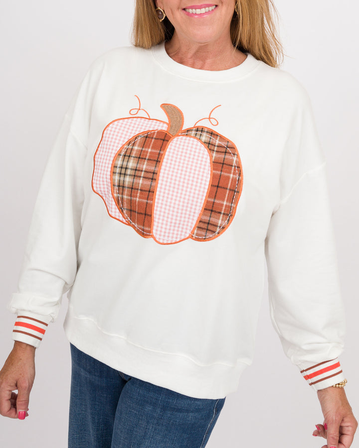 A woman wearing a white crewneck sweatshirt with a large autumn pumpkin graphic on the front. The pumpkin design features orange, brown, and pink plaid and gingham patterns.