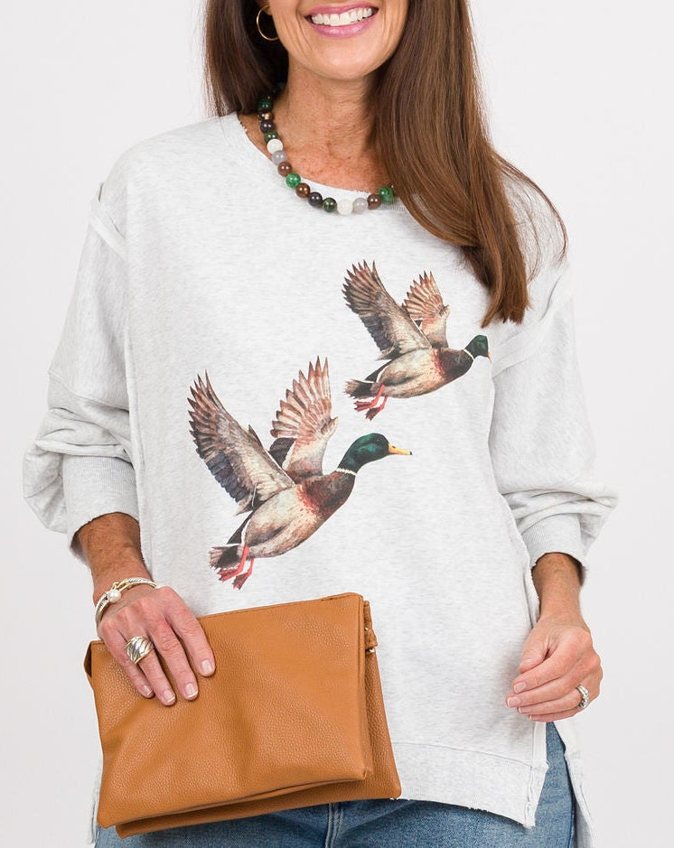 A woman modeling a light gray long-sleeve sweatshirt with a detailed duck graphic on the front, paired with wide-leg blue jeans, tan mule shoes, and a tan clutch bag, set against a plain white studio background.