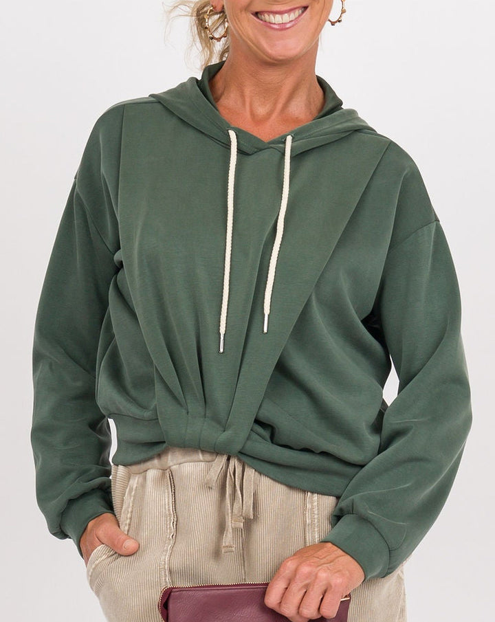A woman modeling a dark green cropped hoodie with white drawstrings, paired with wide-leg beige linen pants and a small burgundy clutch, against a plain white studio background.