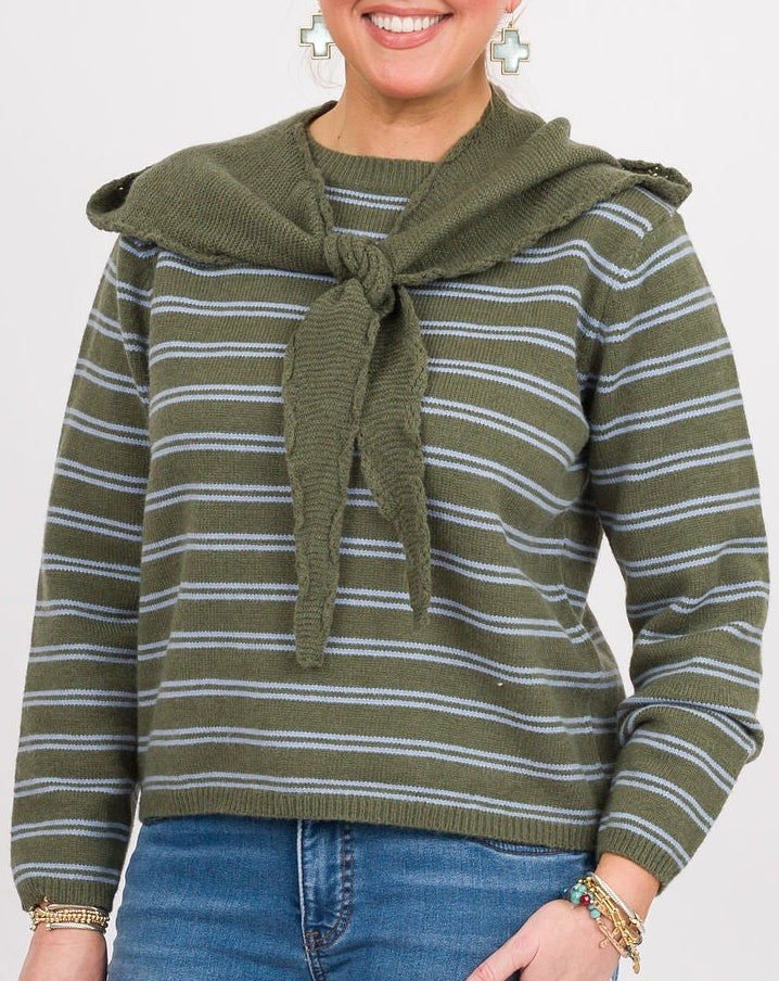 A woman modeling a green and white striped knit sweater with a tie-neck detail, paired with blue jeans and brown clogs, against a plain light gray studio background.