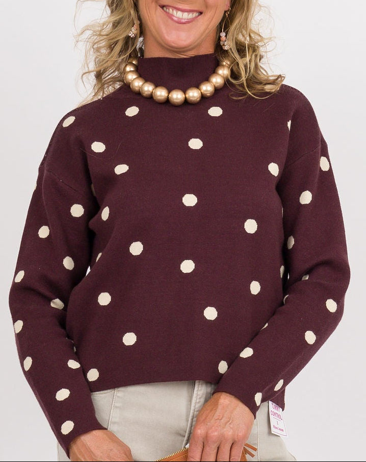 A woman modeling a dark burgundy mock-neck sweater with white polka dots, paired with wide-leg beige pants, brown loafers, and holding a brown clutch against a plain white studio background.