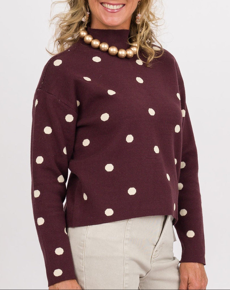 A woman modeling a burgundy mock-neck sweater with white polka dots, paired with light beige wide-leg jeans, brown and cream platform shoes, and holding a brown woven clutch against a plain white studio background.