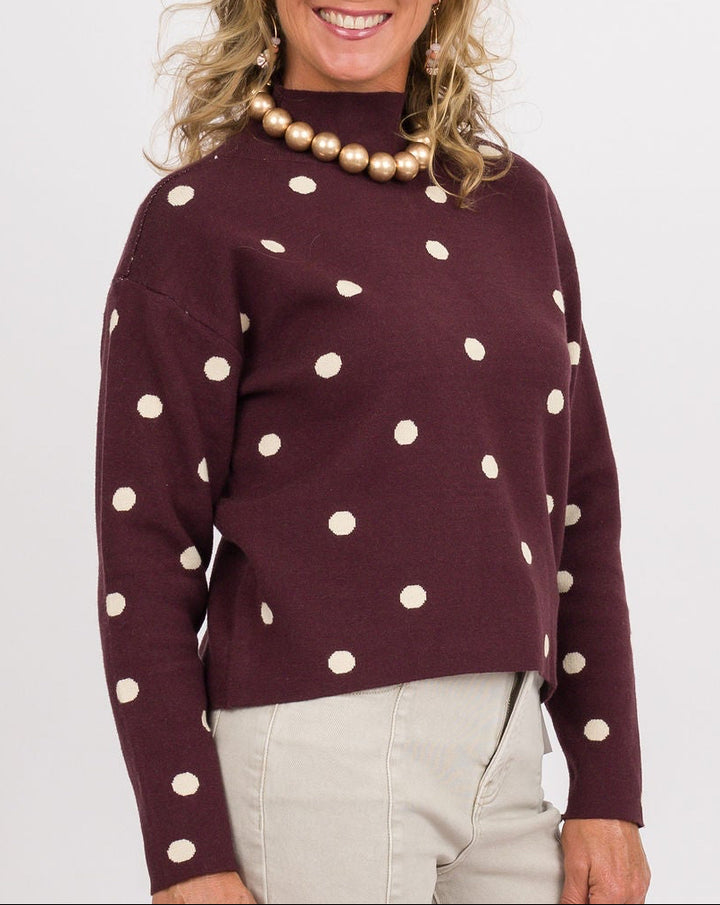 A woman modeling a burgundy mock-neck sweater with white polka dots, paired with light beige wide-leg jeans, brown and cream platform shoes, and holding a brown woven clutch against a plain white studio background.