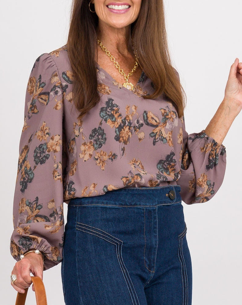 A woman modeling a long-sleeve floral blouse and dark blue wide-leg jeans, holding a brown woven handbag against a plain light gray studio background.