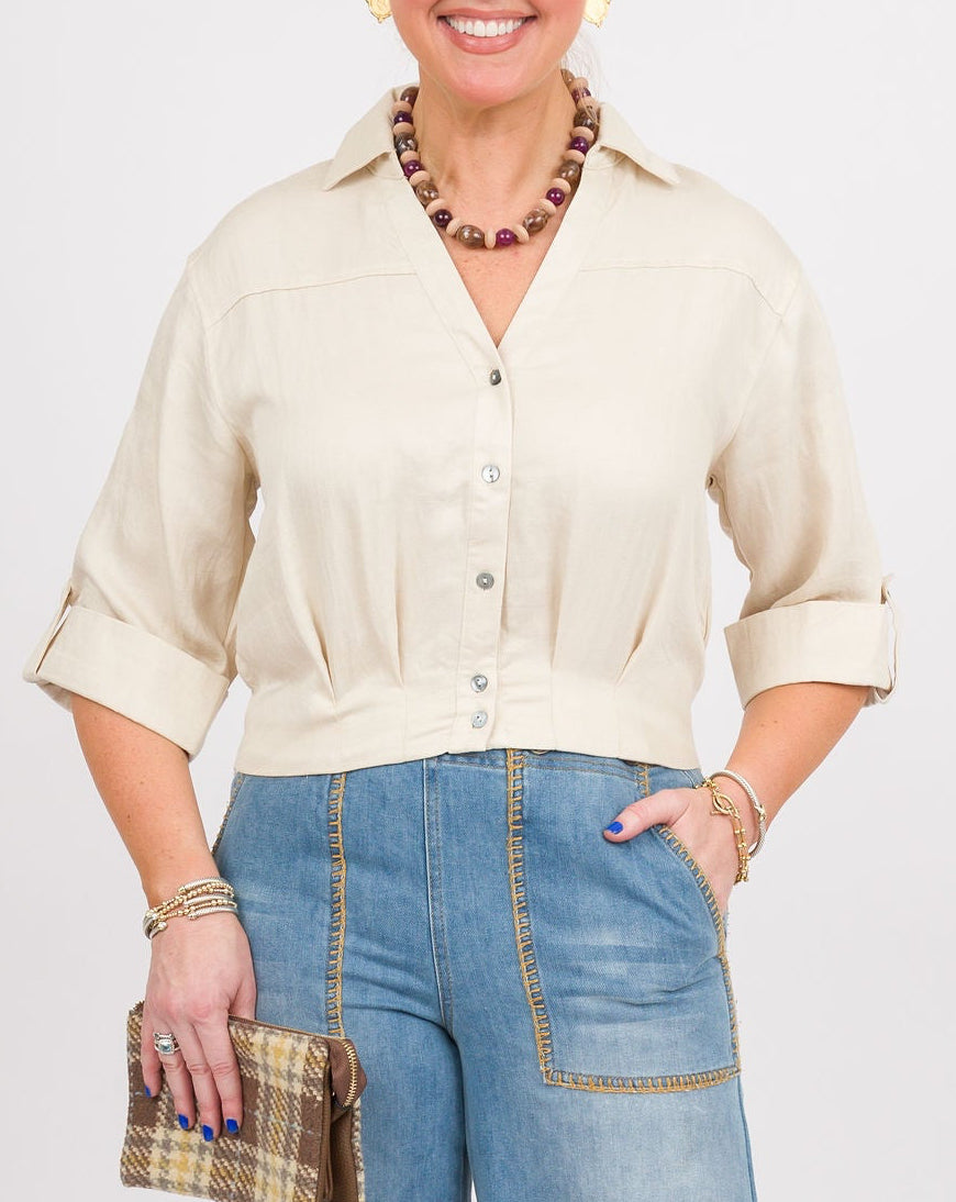 A woman modeling a cream-colored short-sleeve button-up shirt and wide-leg blue denim pants, holding a small plaid clutch and wearing brown clogs, against a plain light gray studio background.