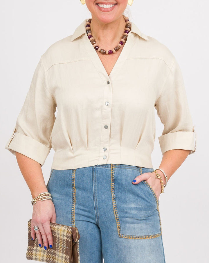 A woman modeling a cream-colored short-sleeve button-up shirt and wide-leg blue denim pants, holding a small plaid clutch and wearing brown clogs, against a plain light gray studio background.
