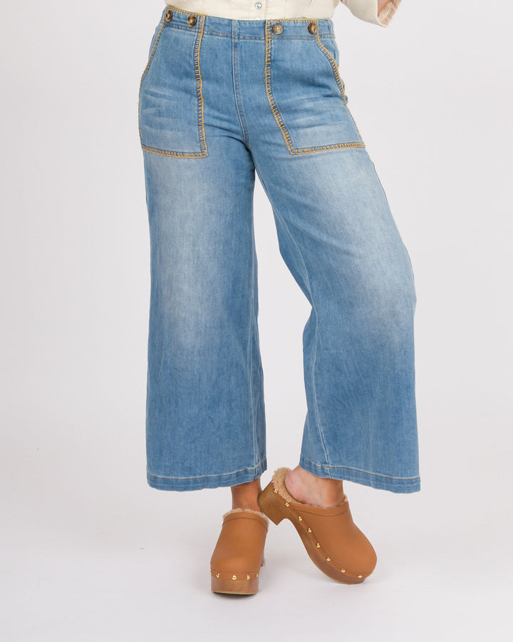 A person wearing light blue wide-leg cropped jeans with contrast tan stitching, paired with tan clogs, photographed against a plain light gray studio background.