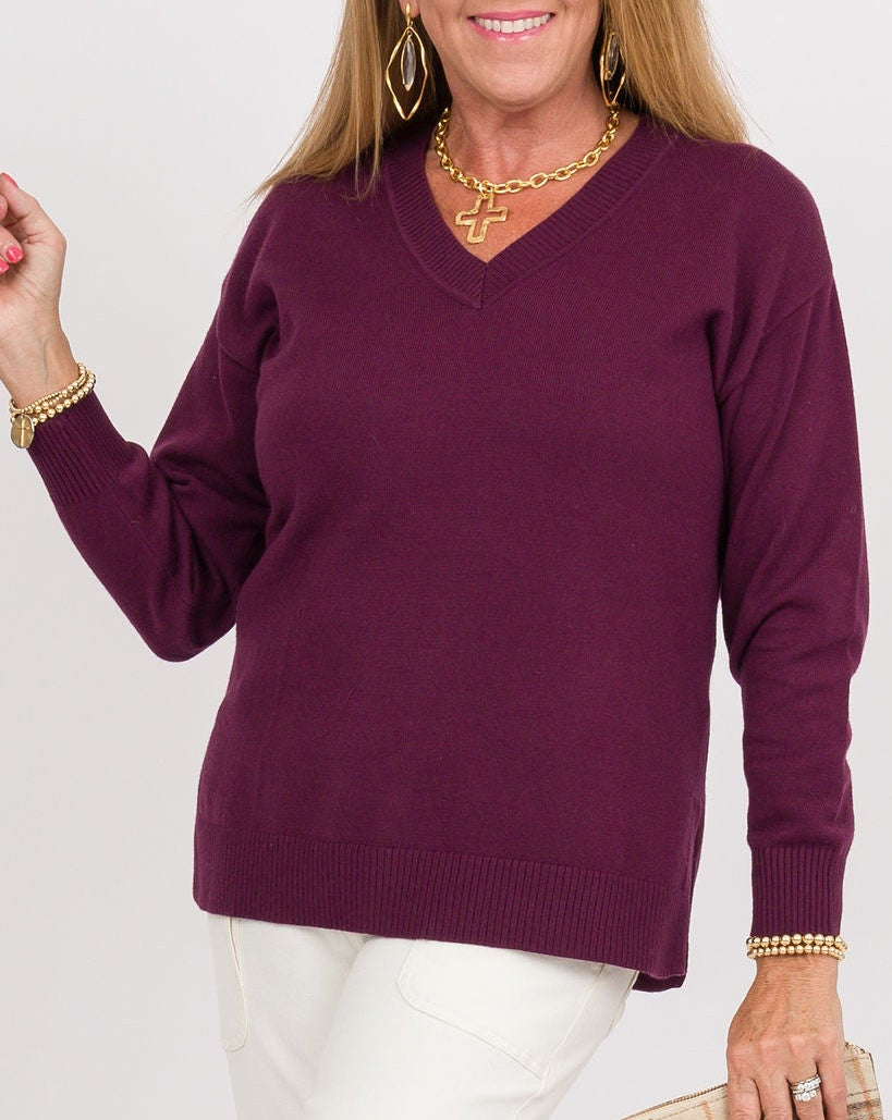 A woman modeling a long-sleeve V-neck knit sweater in deep plum purple, paired with white pants and clear high-heeled sandals, holding a beige plaid clutch against a plain white studio background.