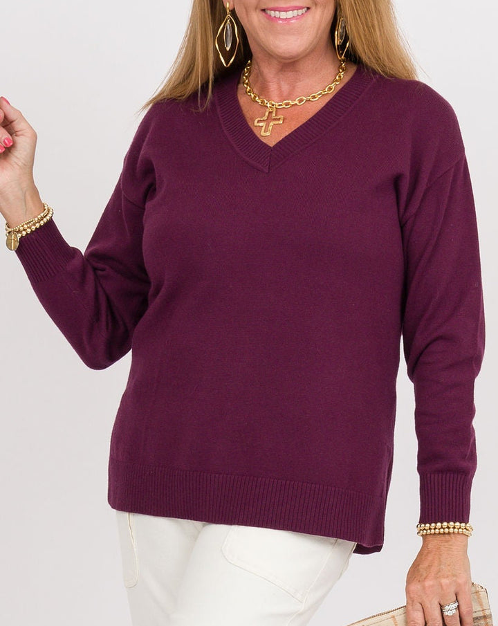 A woman modeling a long-sleeve V-neck knit sweater in deep plum purple, paired with white pants and clear high-heeled sandals, holding a beige plaid clutch against a plain white studio background.