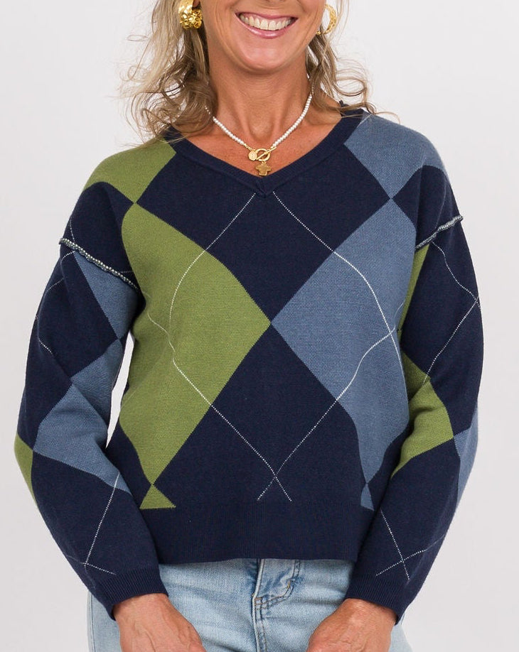 A smiling woman stands against a light gray studio background wearing a navy, blue, and olive green argyle sweater, light-wash wide-leg jeans, and tan shoes, while holding a small beige clutch.