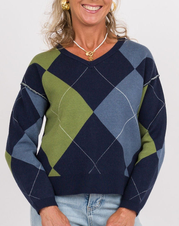 A smiling woman stands against a light gray studio background wearing a navy, blue, and olive green argyle sweater, light-wash wide-leg jeans, and tan shoes, while holding a small beige clutch.